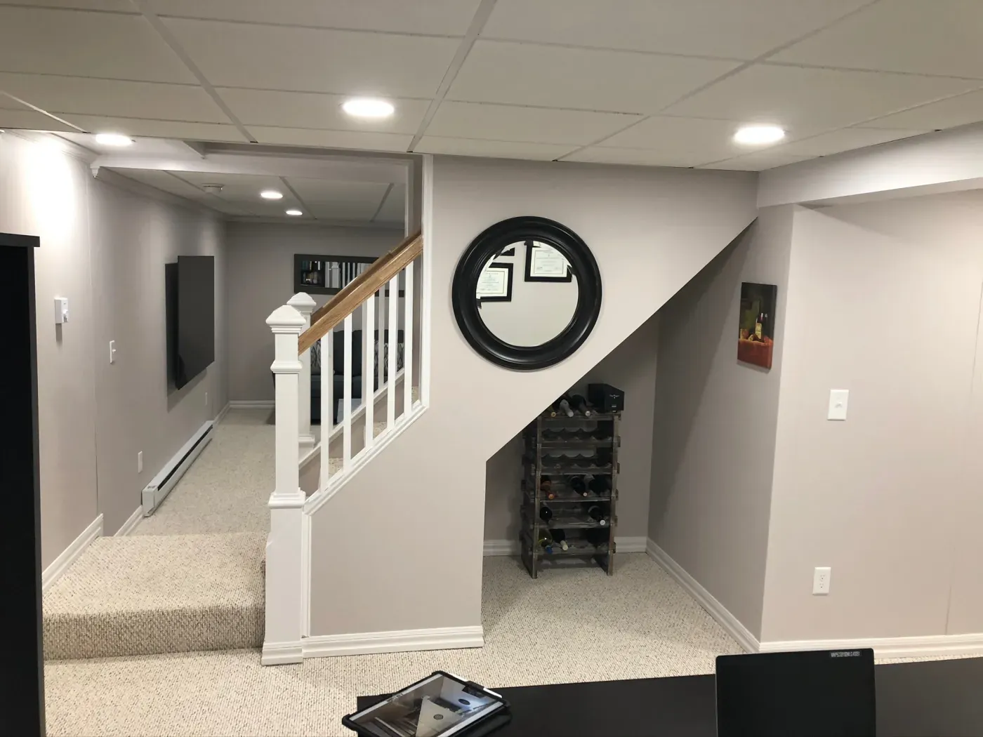 Basement interior with carpeted stairs, white railing, round mirror, and framed certificate. Wine rack under stairs, modern and cozy atmosphere.