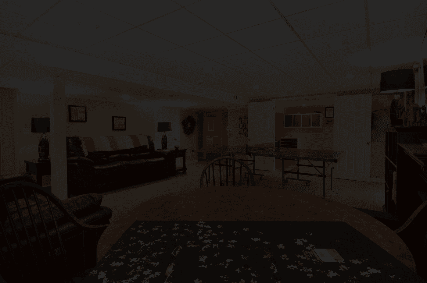 A cozy basement game room with dim lighting, featuring a puzzle on a round wooden table, leather sofa, framed art, and a ping-pong table, exuding a relaxed, inviting atmosphere.