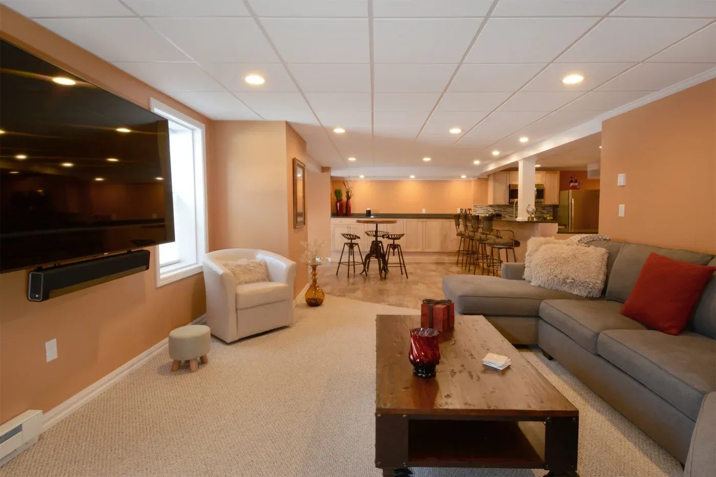 A cozy basement living area with warm orange walls, a large sectional sofa with red and beige cushions, and a flat-screen TV. A wooden coffee table holds decorative vases. In the background, a bar with stools adds a social vibe, while soft lighting enhances the inviting ambiance.