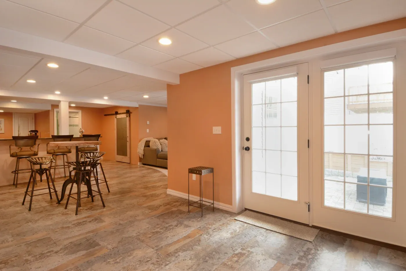 A warm-toned basement with tiled floors, featuring a bar area with brown stools, a cozy couch, and glass doors letting in natural light, creating an inviting atmosphere.