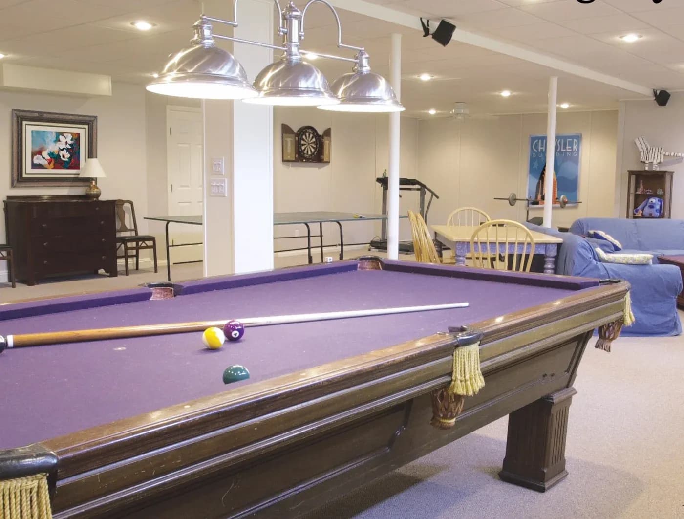 A cozy game room featuring a purple pool table, pool cue, and balls. Nearby are a dartboard, table tennis, blue seating, and wooden chairs.