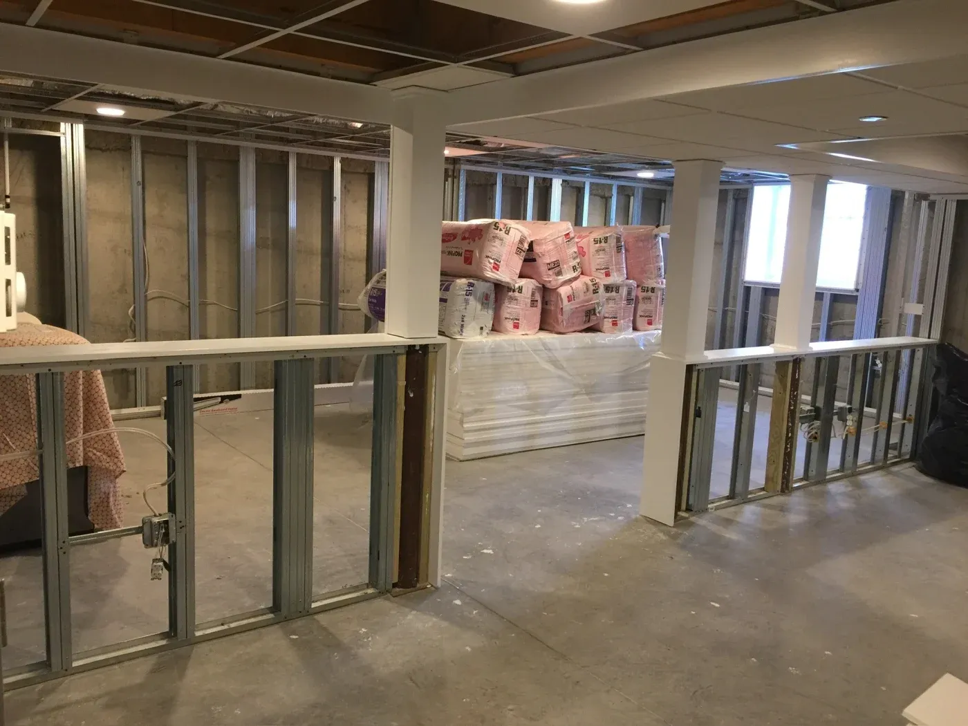 Partially finished basement with exposed metal framing and stacks of insulation materials. Open ceiling with lights, concrete floor, and a covered sofa.