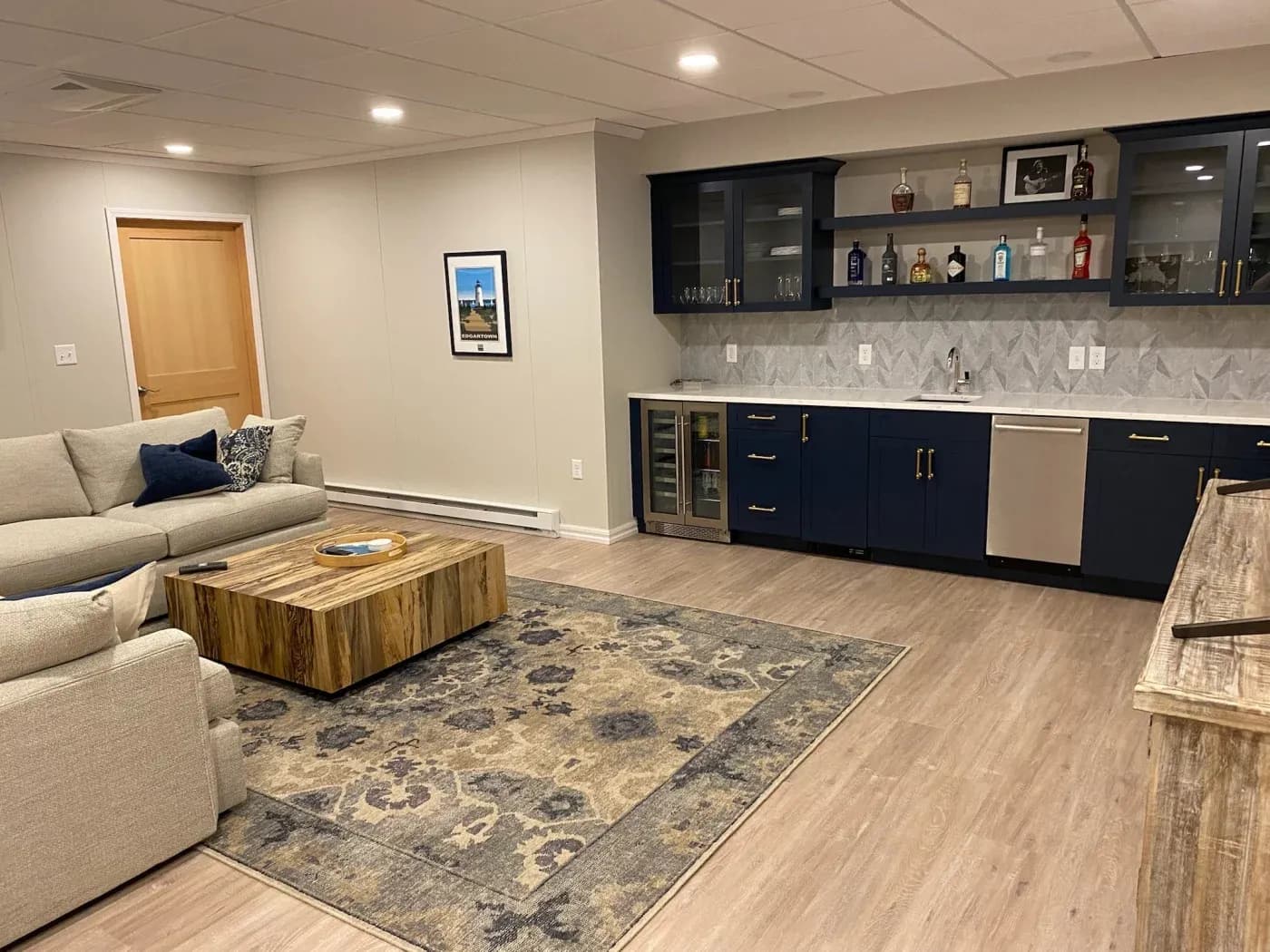 Stylish basement room with navy cabinets, a wet bar, and a large gray rug. Light wood floors, beige couch, and wooden coffee table create a cozy ambiance.