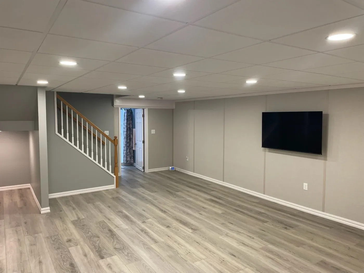 Spacious modern basement with light gray walls, wood flooring, a staircase with a wooden railing, and a wall-mounted TV, conveying a cozy atmosphere.