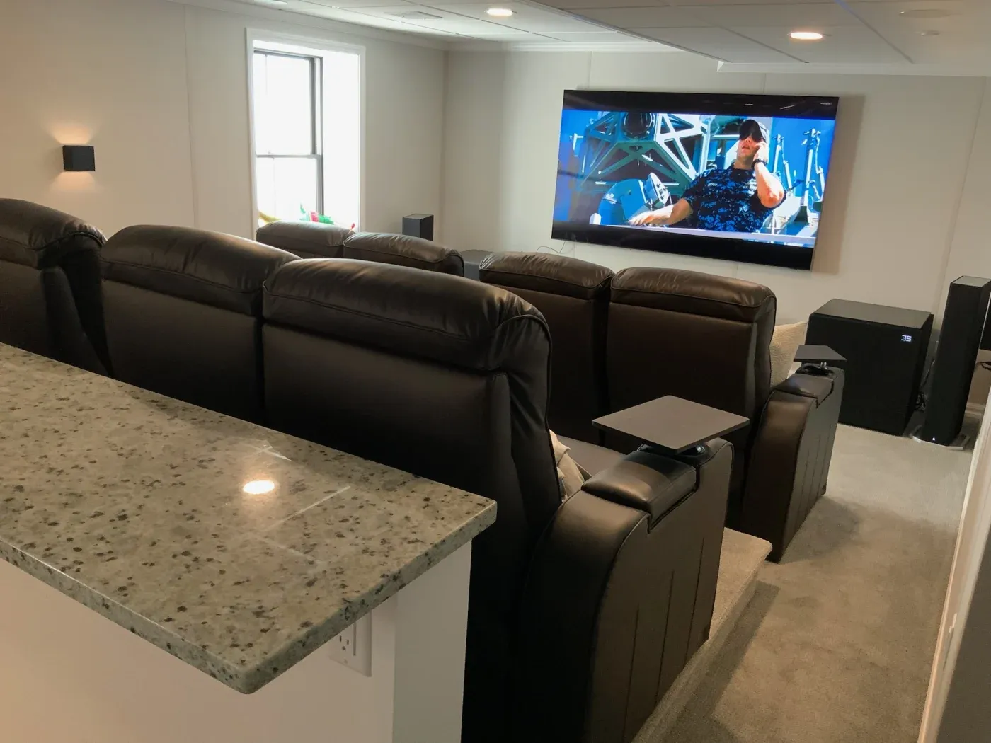 A cozy home theater with leather recliners facing a large wall-mounted screen showing a movie. A granite bar counter is visible in the foreground.