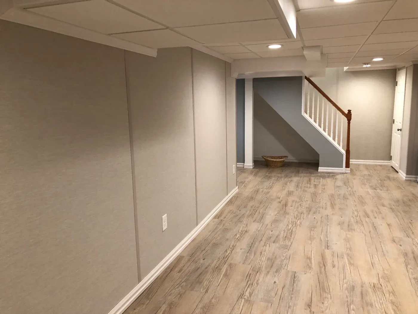 Spacious basement with wood flooring, beige walls, and recessed lighting. Features stairs with a white rail and a wicker basket in the corner. Calm ambiance.