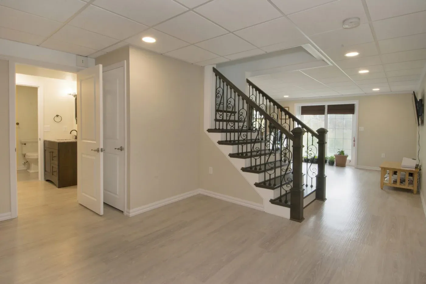 Spacious basement with light wood flooring, white walls, and recessed lighting. Features a staircase with decorative railing, open door to bathroom. Cozy and inviting.